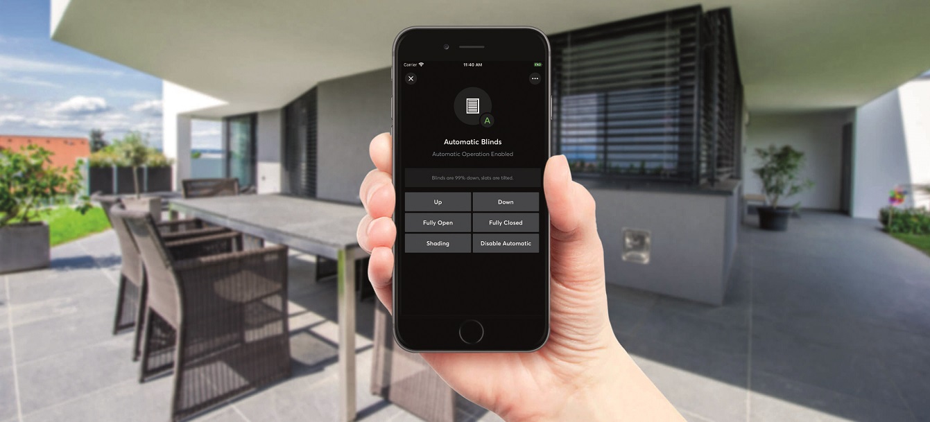 Summert smart home options: Loxone app on mobile phone, in the background the terrace of a family house with the shades down